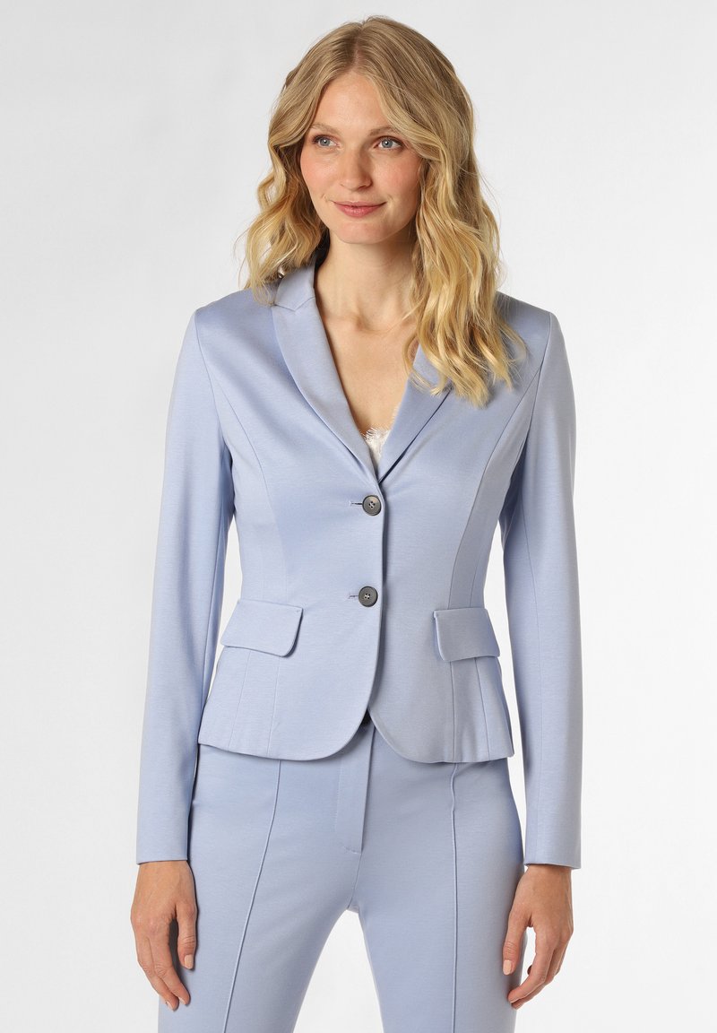 Blonde Frau in einem taillierten hellblauen Blazer und passenden Hosen, die vor einem schlichten weißen Hintergrund steht.