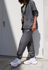 Šedý vzorovaný teplákový komplet s bílými logovými akcenty, doplněný bílými teniskami. Bundu a kalhoty zdobí elastické manžety a lehký design.