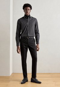 Black striped button-up shirt with long sleeves paired with black tailored pants and black shoes, standing on a wooden floor against a neutral backdrop.