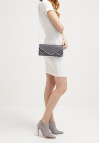 Femme dans une robe blanche ajustée, portant des bottines à talons gris, et tenant un petit sac à main noir pailleté avec une chaîne en argent.