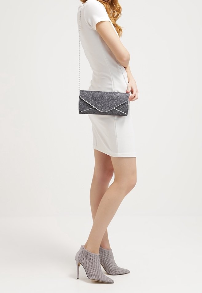 Femme dans une robe blanche ajustée, portant des bottines à talons gris, et tenant un petit sac à main noir pailleté avec une chaîne en argent.