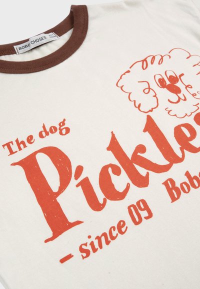 Maglietta color crema con collo marrone, con testo rosso "The dog Pickles - since 09" e un semplice disegno in linea rossa del volto di un cane.