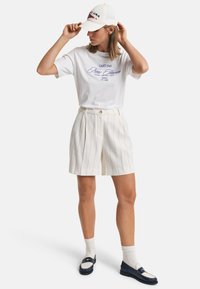 Witte t-shirt met marineblauwe grafische tekst, crèmekleurige gestreepte shorts en witte sokken, gecombineerd met marineblauwe loafers. Een witte pet maakt de outfit compleet.