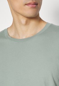 T-shirt en coton vert clair avec un col rond. Texture lisse, manches courtes et coupe ajustée, mettant en valeur le col et la zone des épaules.