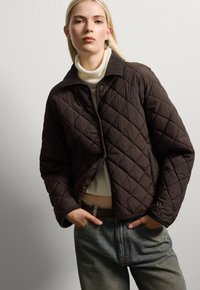 Braune gesteppte Jacke mit Kragen, die ein Rautenmuster und einen Druckknopfverschluss aufweist, getragen über einem hellen Rollkragenpullover und lockeren Jeans.