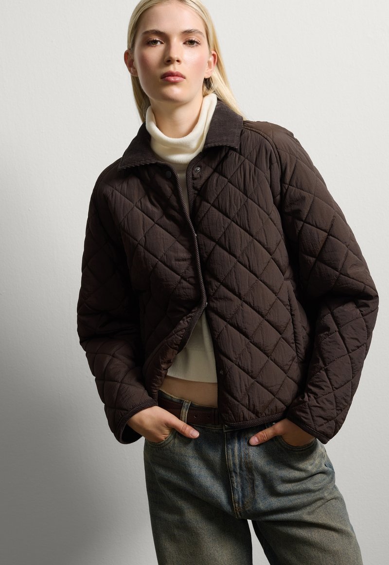 Braune gesteppte Jacke mit Kragen, die ein Rautenmuster und einen Druckknopfverschluss aufweist, getragen über einem hellen Rollkragenpullover und lockeren Jeans.