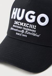 Cappellino da baseball nero con testo ricamato bianco: "HUGO," numeri romani "MCMXCIII," informazioni sul marchio e "Dal 1993."