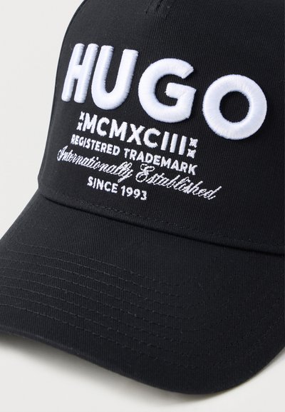 Casquette de baseball noire avec du texte brodé blanc : "HUGO," chiffres romains "MCMXCIII," informations sur la marque, et "Depuis 1993."