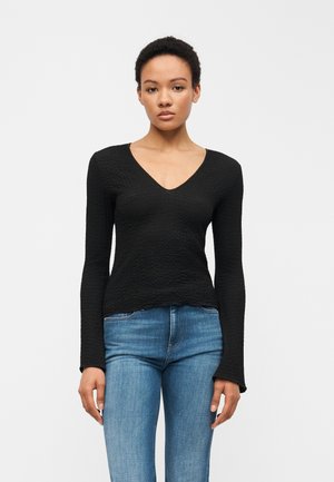 Kobieta ubrana w dopasowaną, czarną, teksturowaną bluzkę z długim rękawem i dekoltem w kształcie litery V oraz niebieskie dżinsy, stojąca na jasnym, jednolitym tle.