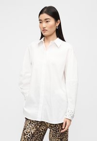 Λευκό βαμβακερό πουκάμισο με κουμπιά και κλασικό γιακά. Διαθέτει διακοσμητικά μανίκια και άνετη εφαρμογή. Συνδυάζεται με παντελόνια με λεοπάρ σχέδιο.