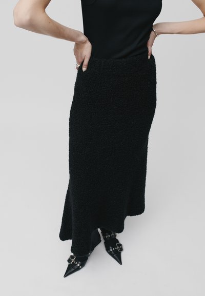 Jupe longue noire en tissu texturé et duveteux avec une taille ajustée. Associée à des chaussures noires à talons pointus avec des accents bouclés.