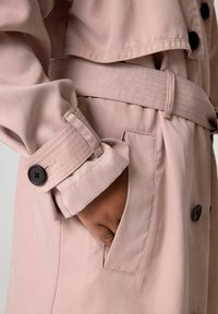 Ljusrosa trenchcoat med bälte vid midjan, stora knappar och en ficka. Tyget verkar slätt med en lätt lyster.