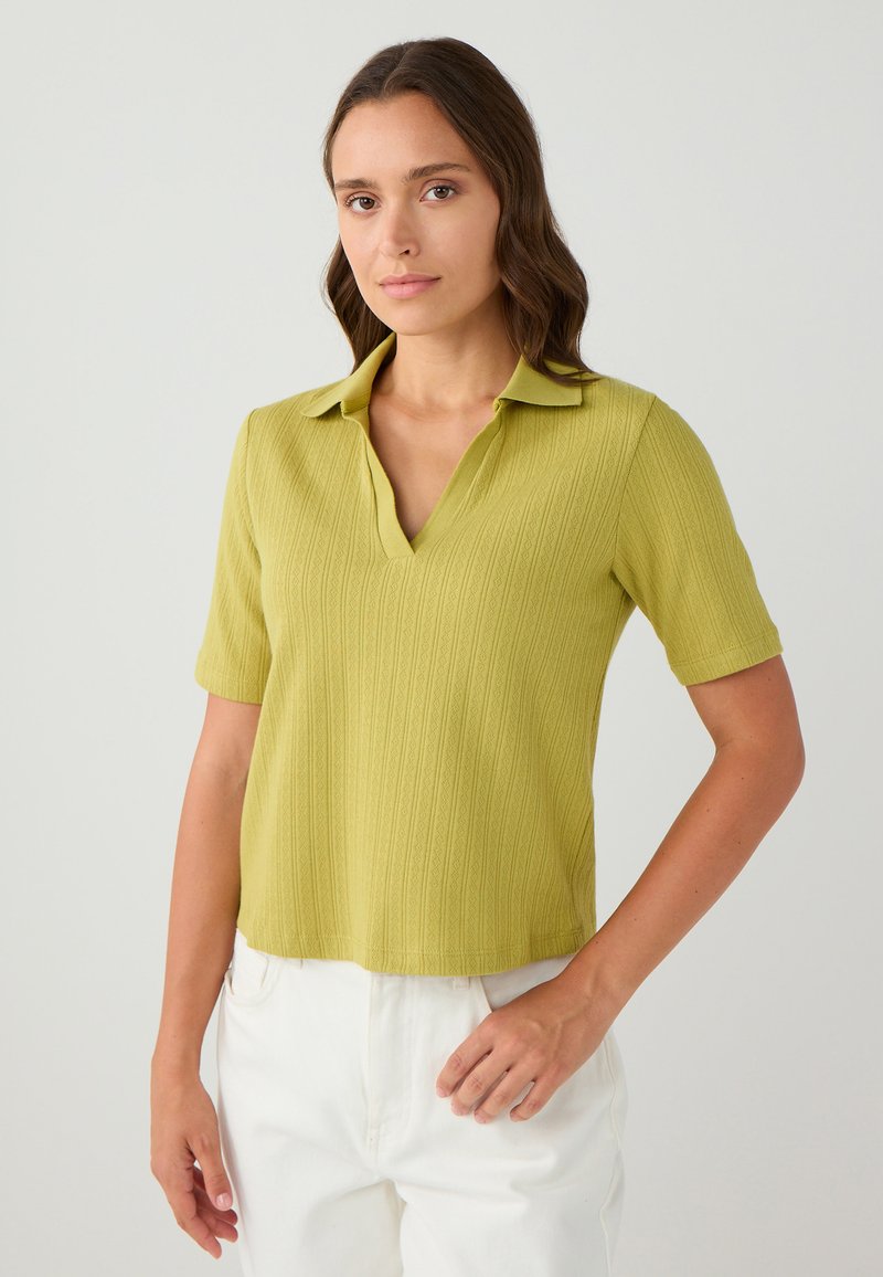 Kurzärmlige, kragenlose Bluse aus hellgrünem Rippstoff. V-Ausschnitt-Design mit dezentem Muster. Kombiniert mit weißen Hosen.