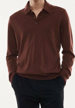 Homme portant un polo en maille brun foncé à manches longues avec col et deux boutons, les mains dans les poches, associé à un pantalon noir.