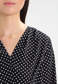 Top in tessuto nero con scollo a V, caratterizzato da un motivo a pois bianchi. Texture morbida, leggera lucentezza e vestibilità ampia.