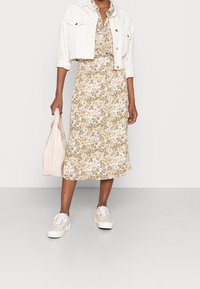 Robe midi à motifs floraux beige, marron et vert ; veste courte en denim blanc ; baskets crème ; tenant un sac fourre-tout de couleur claire.