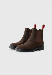 Bottines Chelsea en cuir marron avec des panneaux latéraux élastiques, semelle en caoutchouc noir et tirettes rouges au talon. Texture lisse avec bout arrondi.