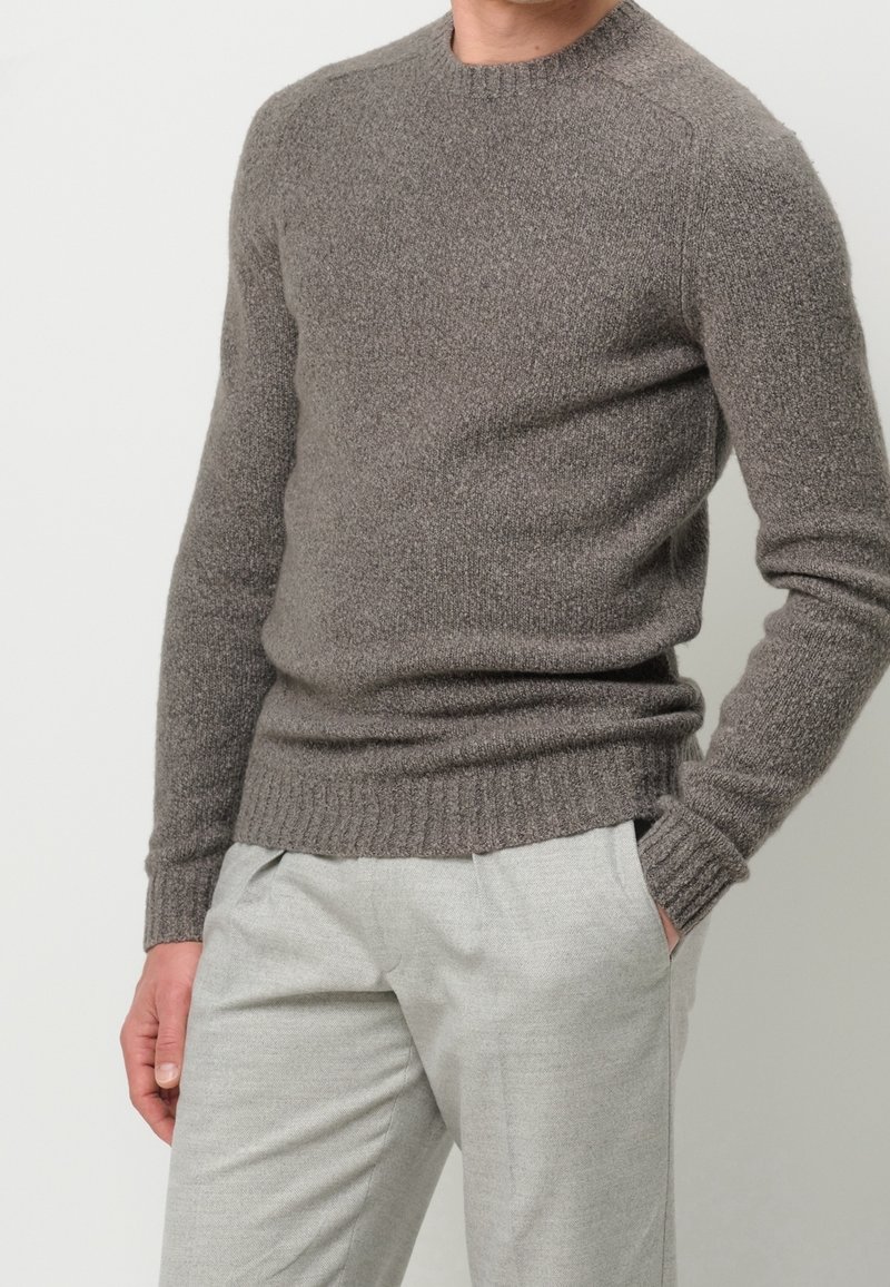Homme portant un pull en maille gris ajusté à manches longues avec poignets et ourlet côtelés, associé à un pantalon gris clair. Une main dans la poche.
