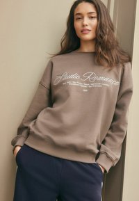 Mladá žena s dlouhými vlasy, oblečená v taupe mikině "Studio Romance" a námořnických kalhotách, opírá se o béžovou zeď a má ruce v kapsách.