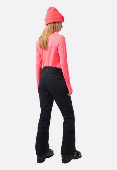 Langärmliges, leuchtend pinkes Oberteil, kombiniert mit schwarzen Skihosen mit Reißverschlüssen an den Knöcheln. Das Modell trägt eine pinke Strickmütze und schwarze Stiefel.
