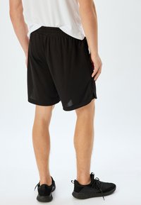 Sorte træningsshorts lavet af letvægtsmateriale, med en elastisk talje, sidelommer og en afslappet pasform, båret med sorte sneakers.