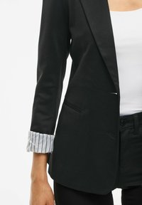 Frau trägt ein schwarzes Blazer mit gestreiften Manschetten, ein weißes Oberteil und schwarze Hosen vor einem weißen Hintergrund, ab Schulter bis Hüfte gezeigt.