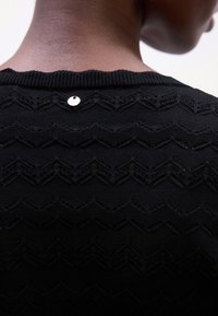 Maglione nero lavorato a maglia con un motivo a zigzag, scollatura a costine e un piccolo bottone d'accento argentato sulla schiena. Texture morbida.