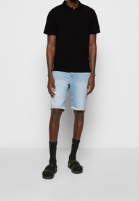 Czarna koszulka polo, jasnoniebieskie dżinsowe szorty podwinięte na mankietach, czarne skarpety i sandały. Prosty, codzienny strój z kontrastowymi kolorami i teksturami.