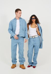 Jachetă din denim de culoare albastru deschis, cu logo brodat, blugi lejeri asortați, topuri albe fără mâneci și curea cu buline; modelul poartă încălțăminte în nuanțe de maro și roșu.