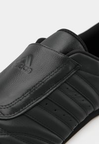 Μαύρο δερμάτινο παπούτσι με υφασμάτινη επιφάνεια, διαθέτοντας βέλκρο, τρυπητά σχέδια στα πλάγια και ένα διακριτικό λογότυπο στο πάνω μέρος.