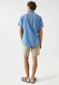 Camisa de manga corta azul claro con collar, hecha de tela de mezclilla; combinada con pantalones cortos beige y sandalias. Vista trasera destacando la longitud de la camisa.