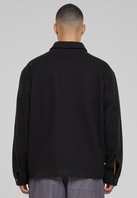 Black oversized jacket with a collar, long sleeves, side slits, and buttoned cuffs. The fabric appears smooth and lightweight.