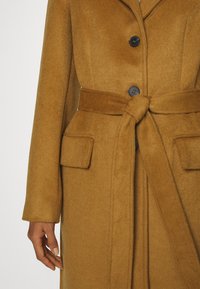 Kamelfärgad ullrock med bältad midja, stora sidfickor och tre svarta knappar. Har en krage med notch och mjuk textur.