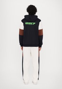 Navy hoodie with green EA7 logo on the back, white and brown accents, paired with cream pants featuring navy side stripes. Soft, textured fabric.