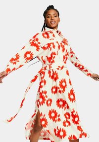 Vestido floral en crema claro con grandes estampados de flores rojas, mangas largas, cuello con solapa, cinturón en la cintura y detalles de abertura lateral.