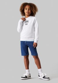 Felpa bianca con testo nero; pantaloni corti in denim blu; sneakers bianche con dettagli neri; capelli ricci; sfondo semplice.