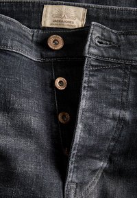 Lähivõte tumehallidest teksapükstest, millel on osaliselt nupud lahti, metallnupud ja kangast silt, millel on kirjutatud "Jack & Jones Jeans Intelligence."