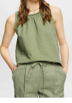 Kvinde iført lyse olivengrøn ærmeløs top og matchende hørbukser med bælte, hænder i lommerne, stående mod ensfarvet baggrund.