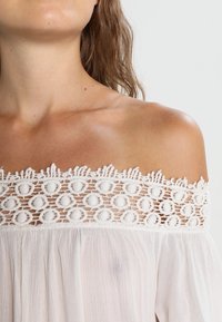 Top blanc à épaules dénudées avec une bordure en dentelle crochetée texturée à motifs ronds, en tissu léger avec un tombé doux.