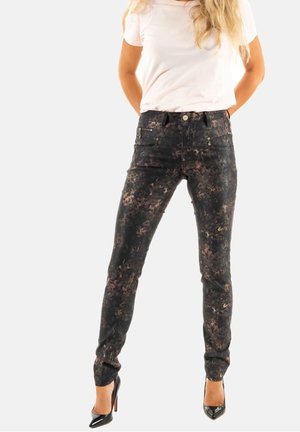 Femme portant un pantalon slim noir et marron à motifs, une chemise blanche à manches courtes et des chaussures à talons noirs, debout, les mains dans le dos.