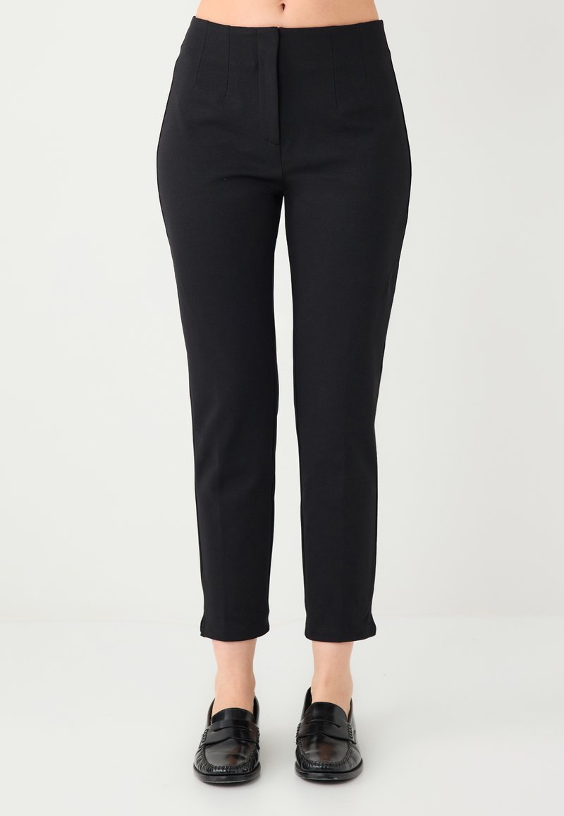 Pantalon noir ajusté avec une coupe slim, présentant une texture lisse, des plis à l'avant et une longueur courte, associé à des mocassins noirs.