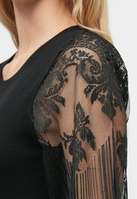 Juodas topas, turintis solidžią medvilnės kūno dalį ir permatomas nėriniuotą rankoves su gėlių raštais ir sviestuvais aplink kraštus.