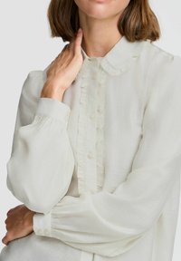Helles Creme-Bluse mit Rüschensaum, Knopfleiste und voluminösen Ärmeln. Weicher Stoff mit gerafften Manschetten und dezentem Glanz.