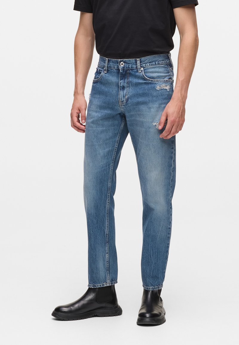 Heledad sinised denim teksad lõdva lõikega, mis sisaldavad kulunud detaile, viit taskut ja nööpkinnitust. Kantud mustade maanduvate saapadega.