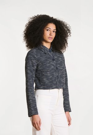 Femme aux cheveux bouclés et volumineux portant une veste courte texturée sombre boutonnée et un pantalon blanc taille haute, se tenant devant un fond uni.