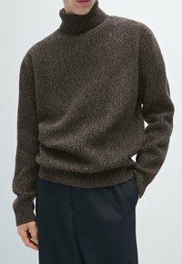 Brązowy sweter dziany z wysokim kołnierzem, z teksturowanym wzorem i luźnym krojem. Połączony z ciemnymi spodniami, ręce w kieszeniach.