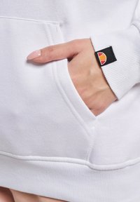 Hånd med manikyrerte negler i frontlomma på en hvit hettegenser med en liten svart etikett som har en rød og oransje logo på mansjetten.
