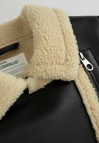 Czarna skórzana kurtka z kremowym kołnierzem z owczej wełny. Zawiera srebrny zamek błyskawiczny oraz widoczny metkę w środku. Teksturowane materiały kontrastują ze sobą w sposób efektowny.