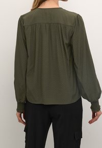 Blouse vert olive avec un motif texturé, coutures froncées aux épaules, poignets élastiqués et coupe décontractée, associée à un pantalon noir.