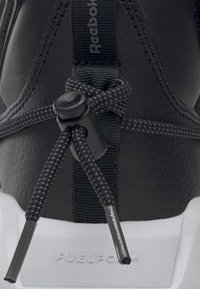 Svart sportskor med texturerat läderöverdrag, elastiska snörningar med en spänne och en vit sula präglad med "FUELFOAM."